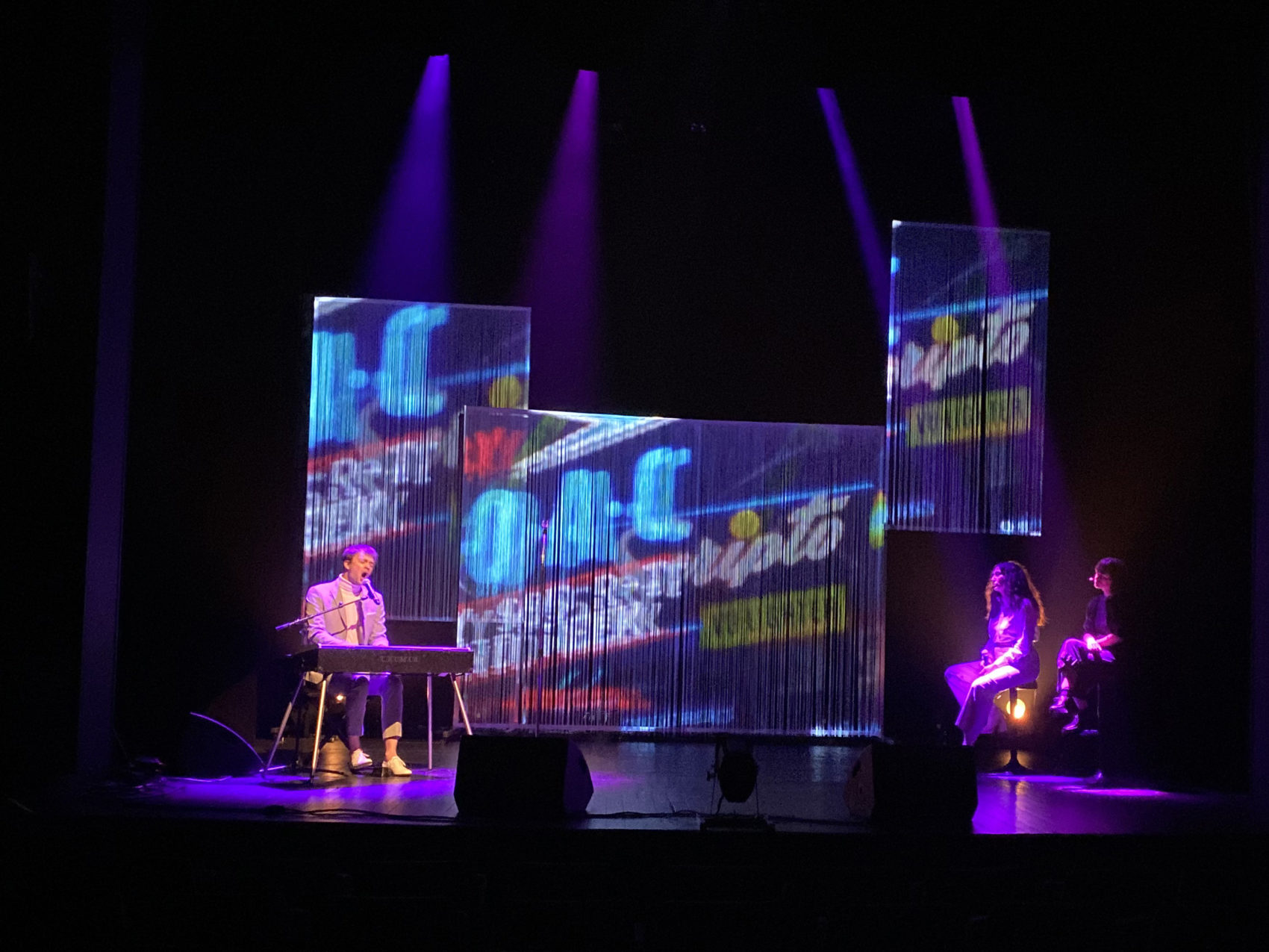 Répétition du spectacle sur scène avec le chanteur à gauche au piano, et deux femmes assises à droite de la scène