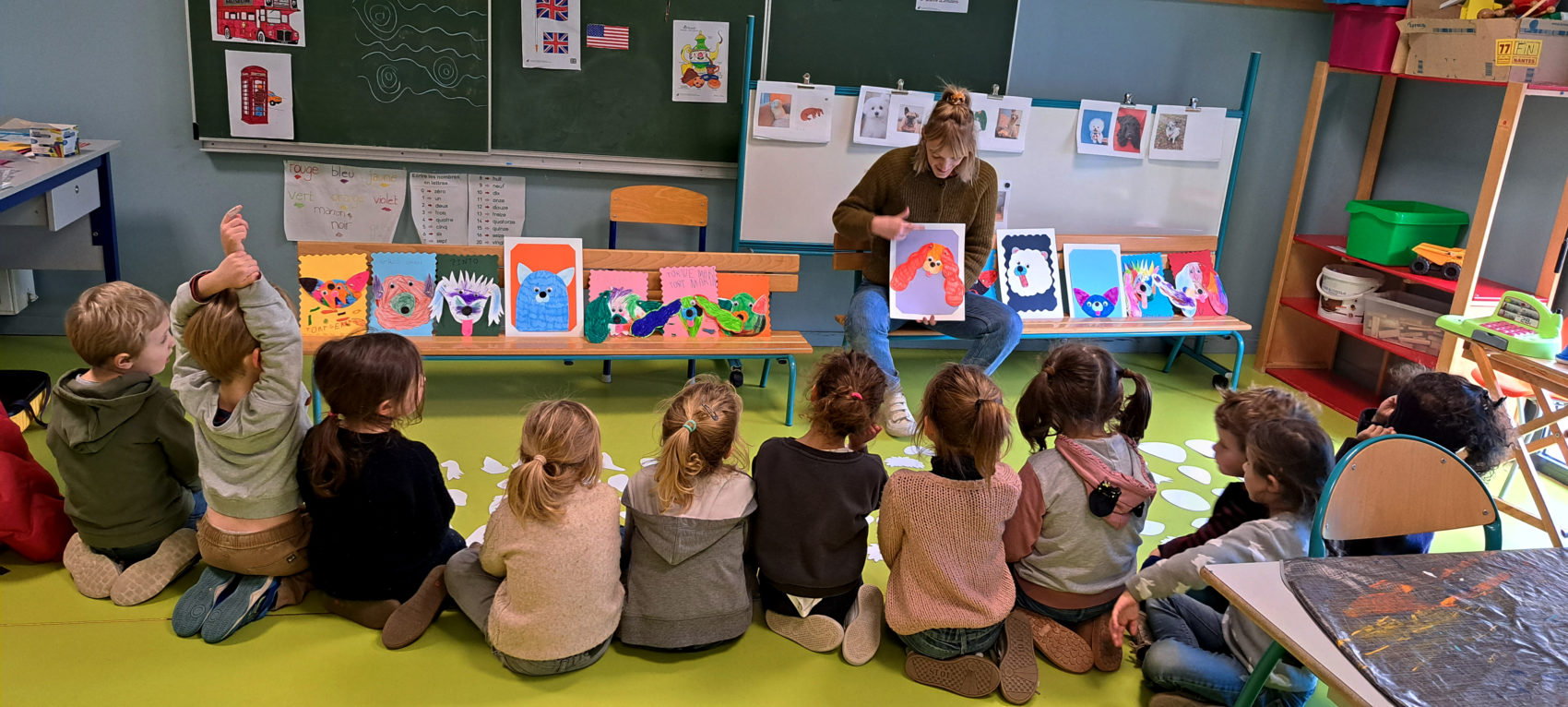 En classe les enfants sont assis de dos devant une femme qui leur présente un dessin de chien