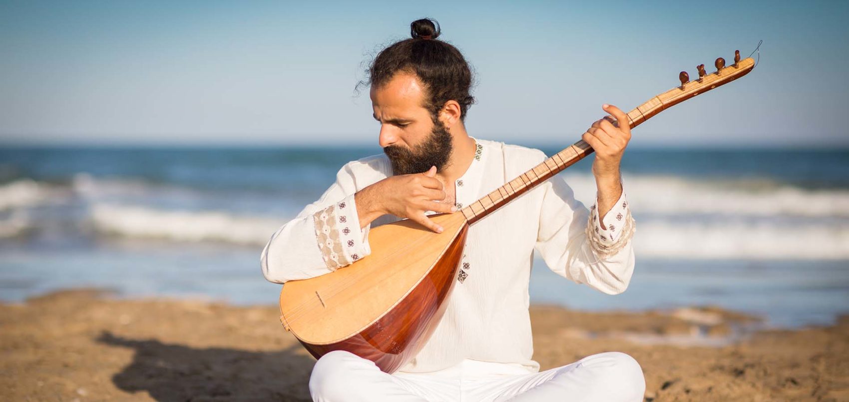 Photo de Rusan Filiztek assis dans le sable, en blanc, jouant de son instrument le oud