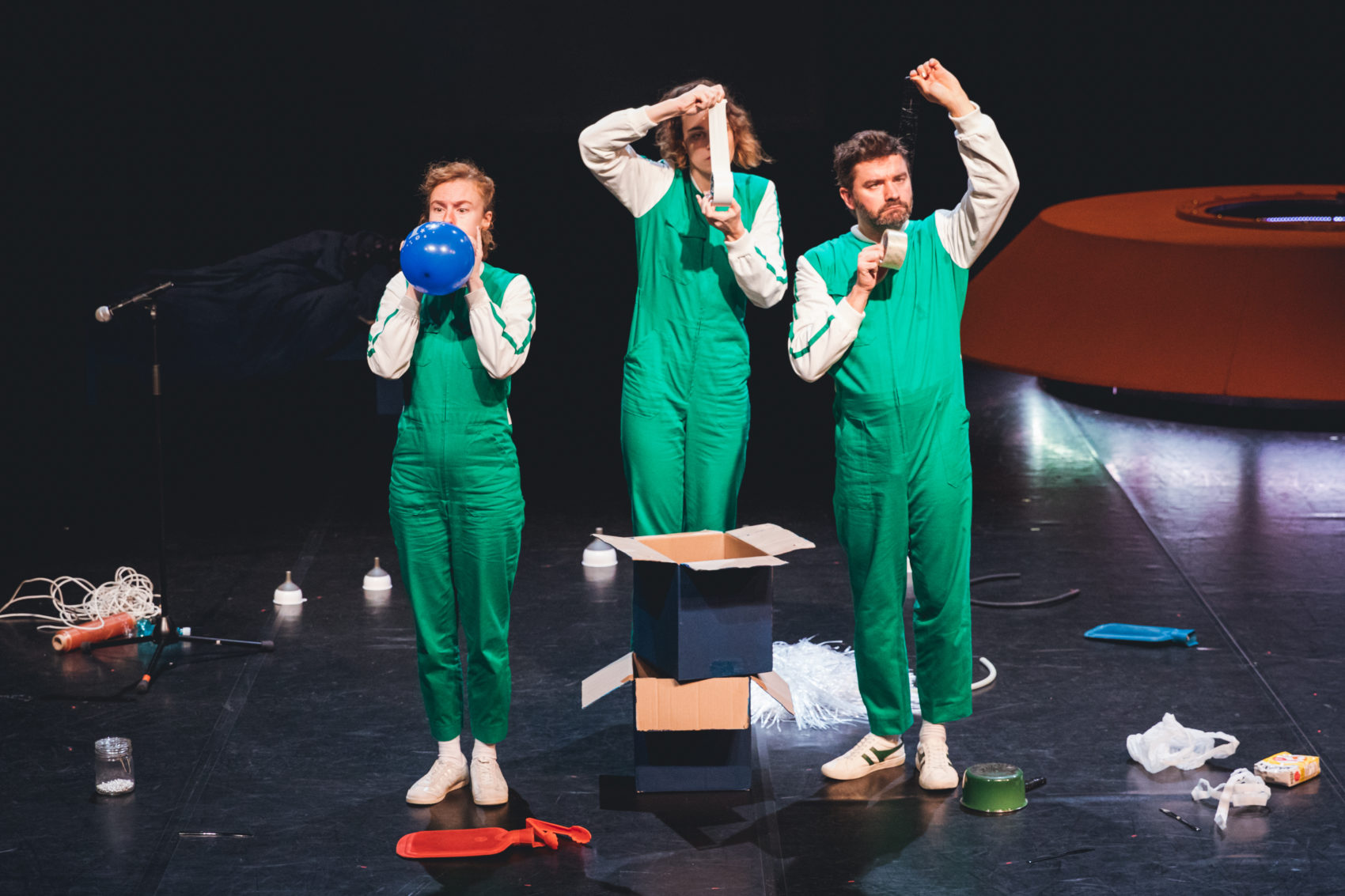 Trois comédiens sur scène habillés en combinaisons verte avec des objets dans les mains