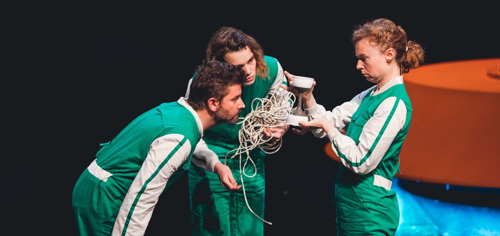 Trois comédiens sur scène en cercle, habillés en combinaisons verte regardent de la corde et un objet avec interrogation 