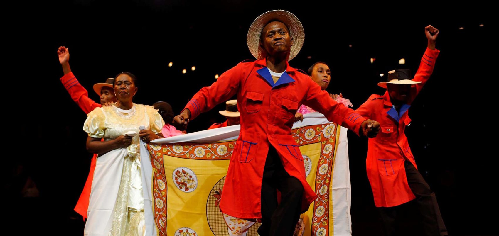 Photo de la troupe qui danse avec un homme en veste traditionnelle rouge au premier plan. 