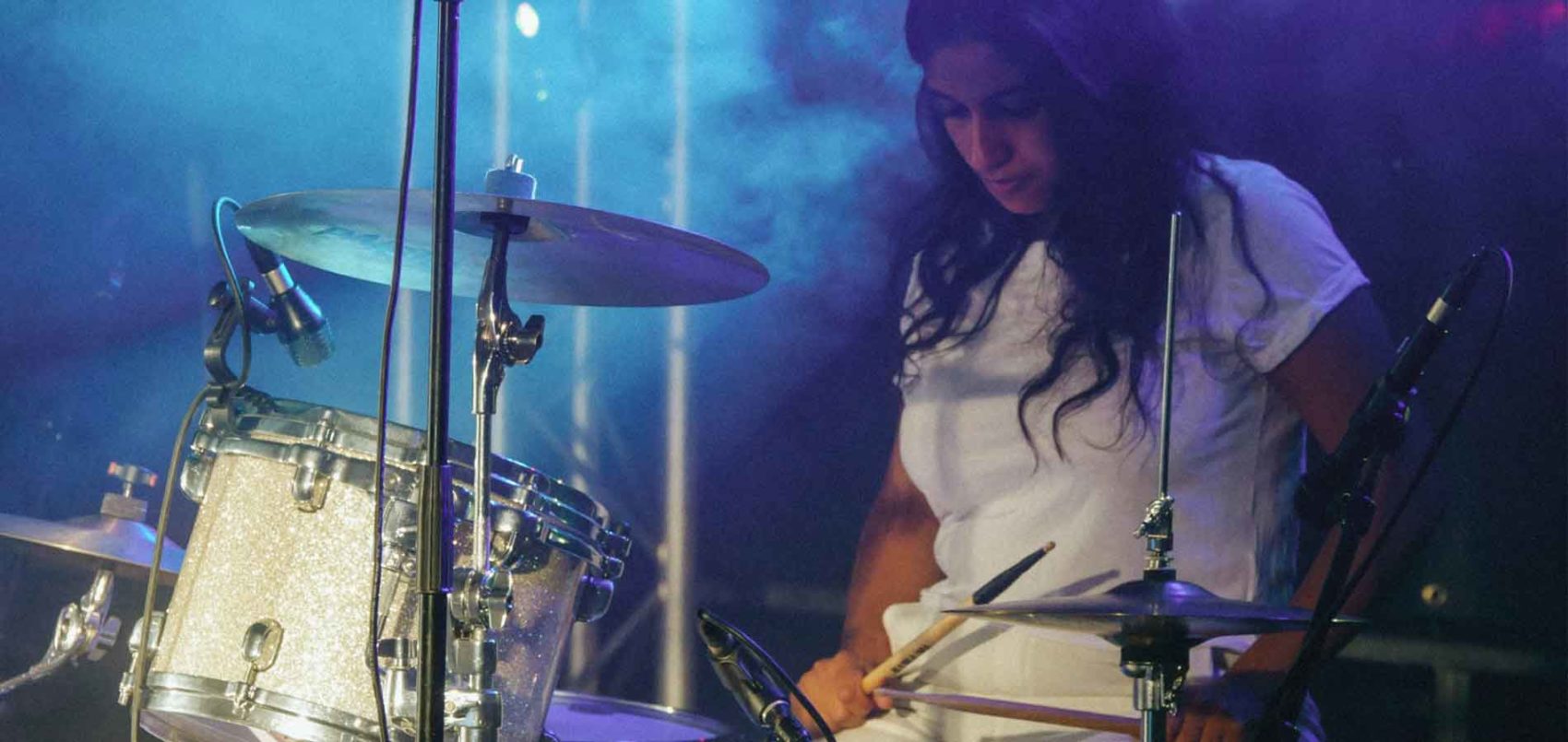 Gaouta, en t-shirt et pantalon blanc qui fait de la batterie sur scène 
