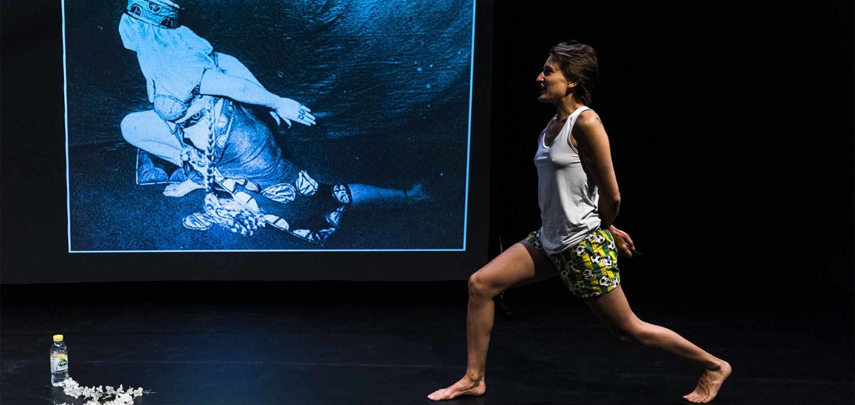 Une femme en débardeur et short qui reprend la même pose que l'image projetée derrière elle