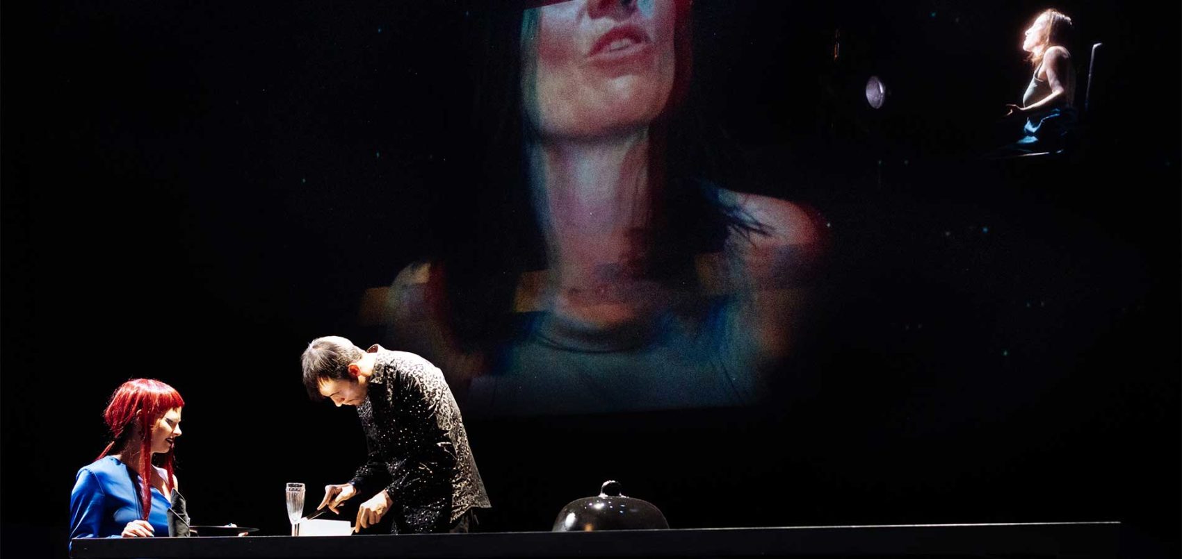 Sur scène, le portrait d'une femme projetée et une femme et un homme face autour d'un bureau