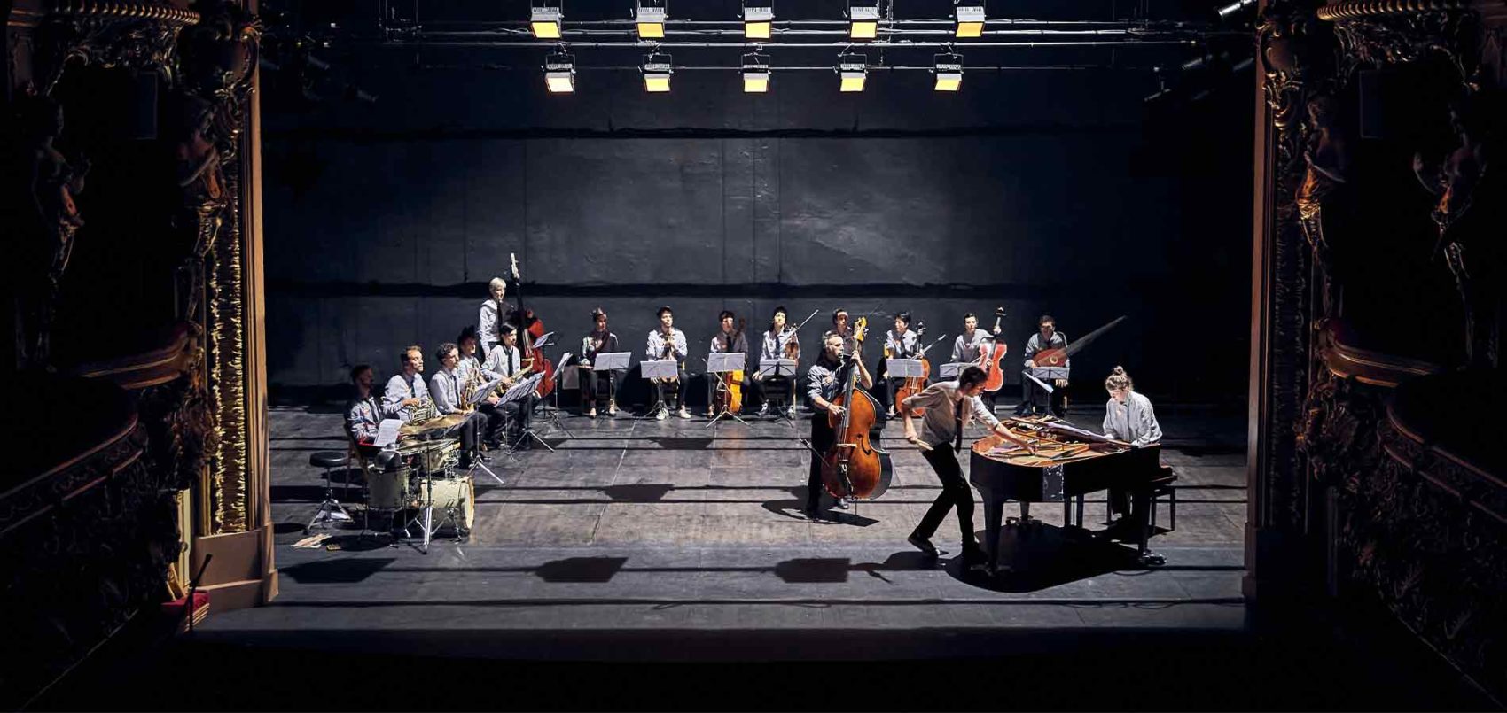Plan d'ensemble de l'orchestre sur la scène. Ils et elles sont en carré et un piano et une contrebasse sont au premier plan à droite