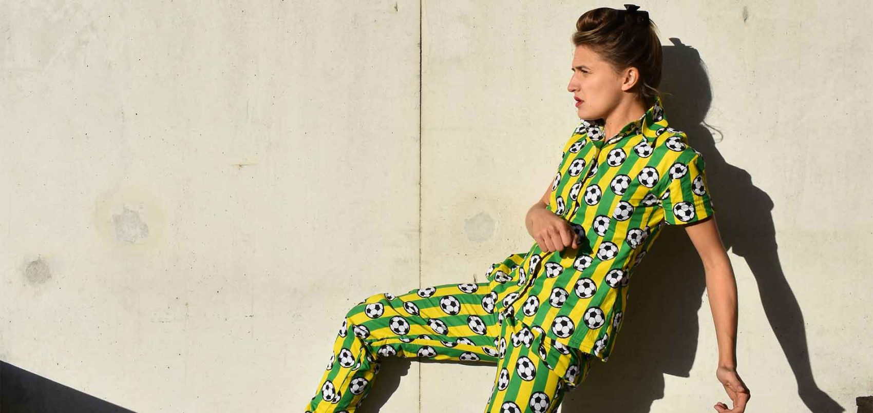 Une femme est en mouvement, de profil et porte un ensemble vert et jaune avec des ballons de foot