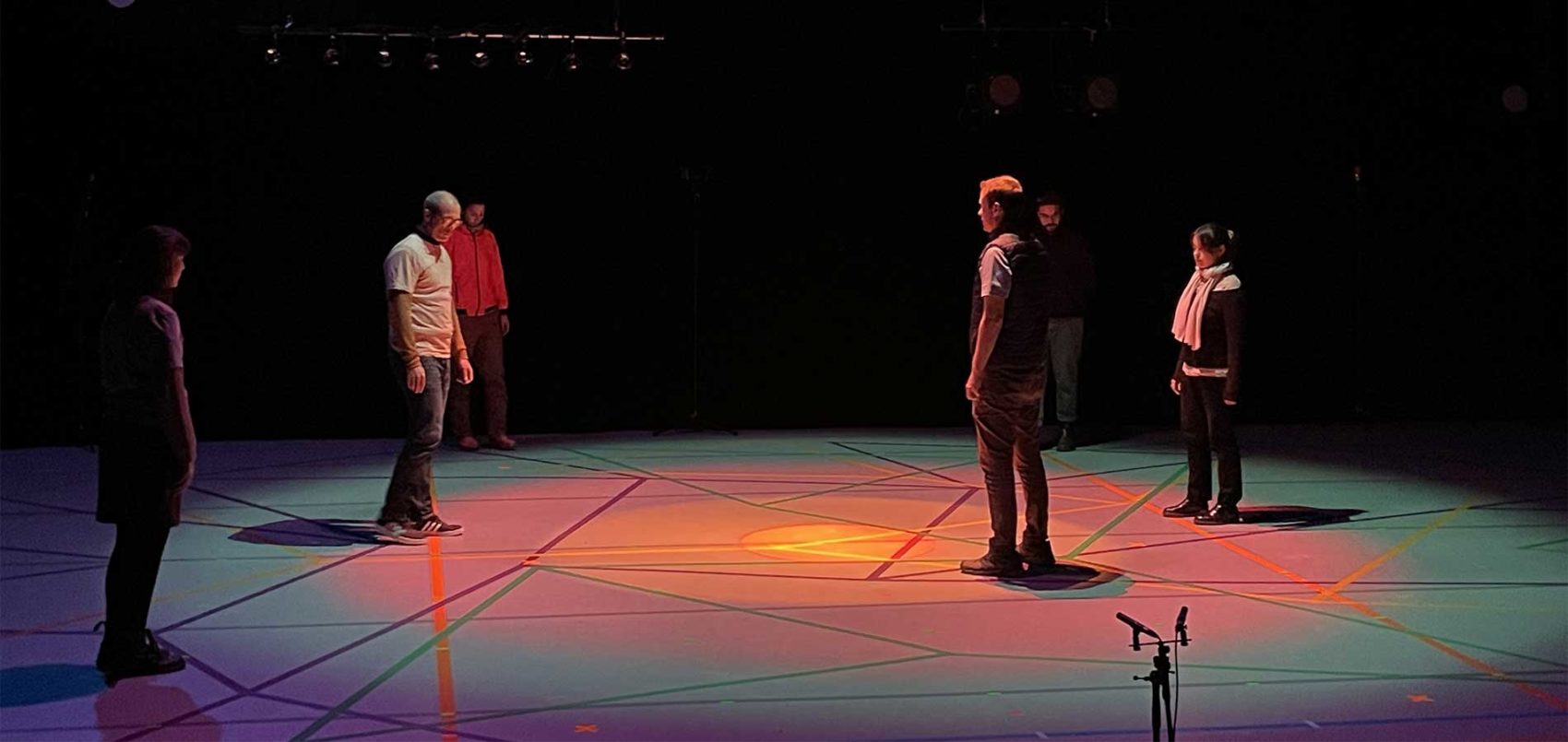 Six personnes sont sur la scène qui est illuminée de plusieurs couleurs. Il y a des grands traits de couleurs au sol