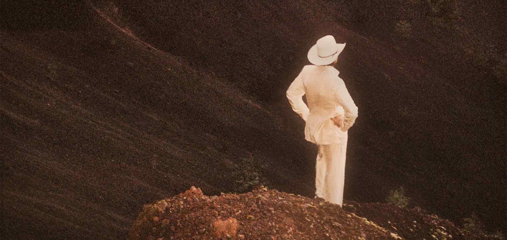Un homme en costume blanc et chapeau de cowboy de dos sur un rocher