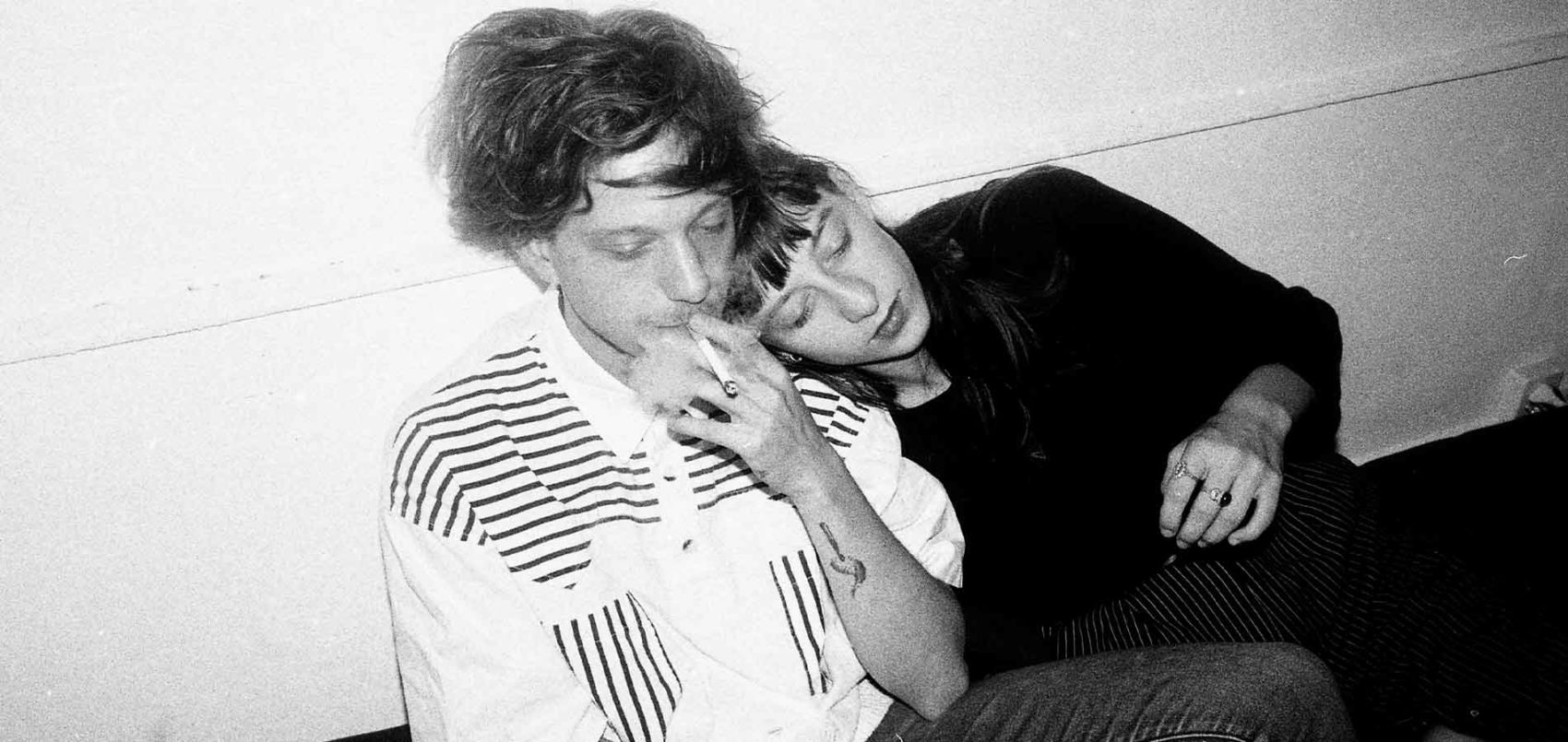 Un homme assis contre un mur qui fume et une femme avec la tête posée contre l'épaule de l'homme 
