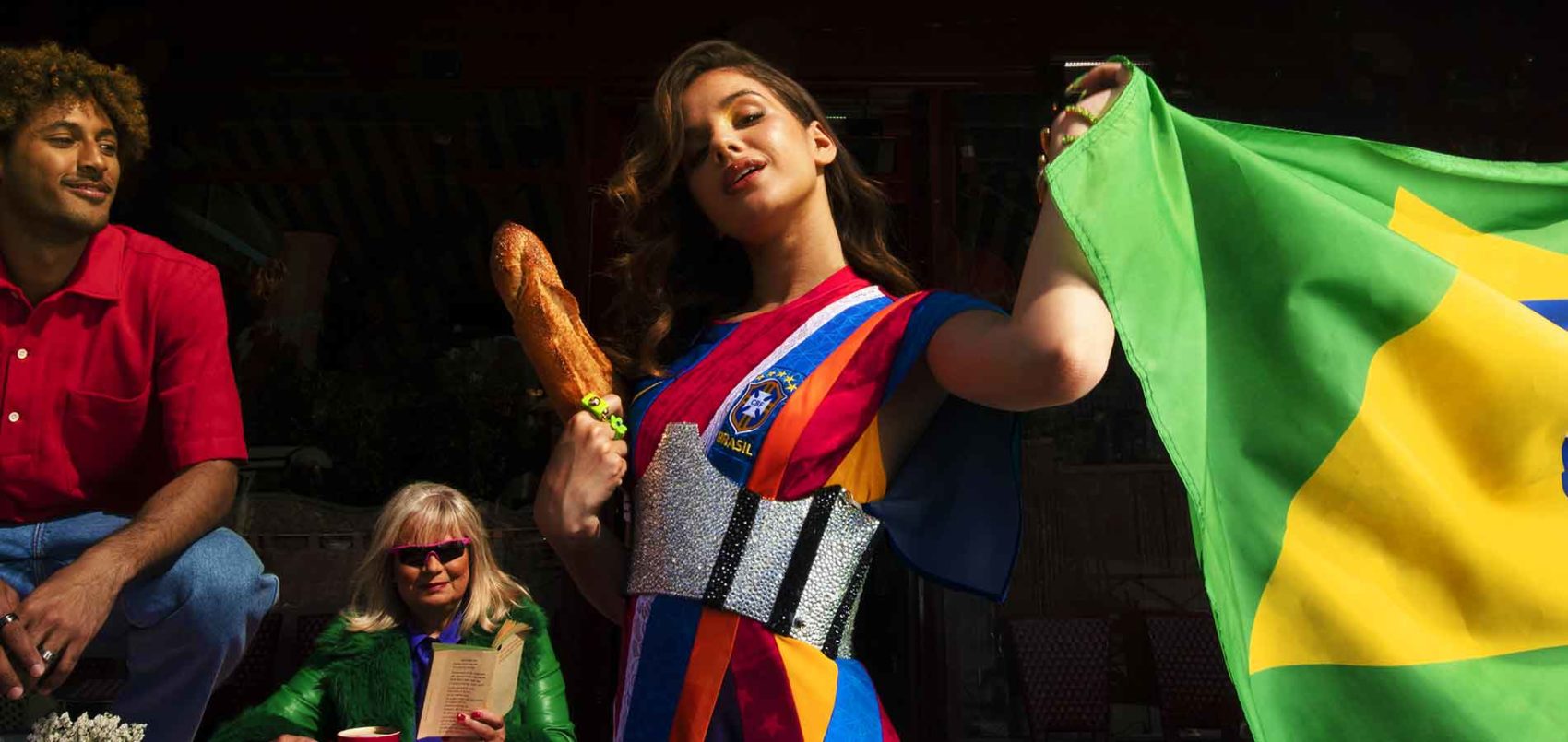 Portrait de Bianca Costa avec un drapeau du Brésil dans la main gauche et une baguette de pain dans la main droite. elle porte un maillot de foot