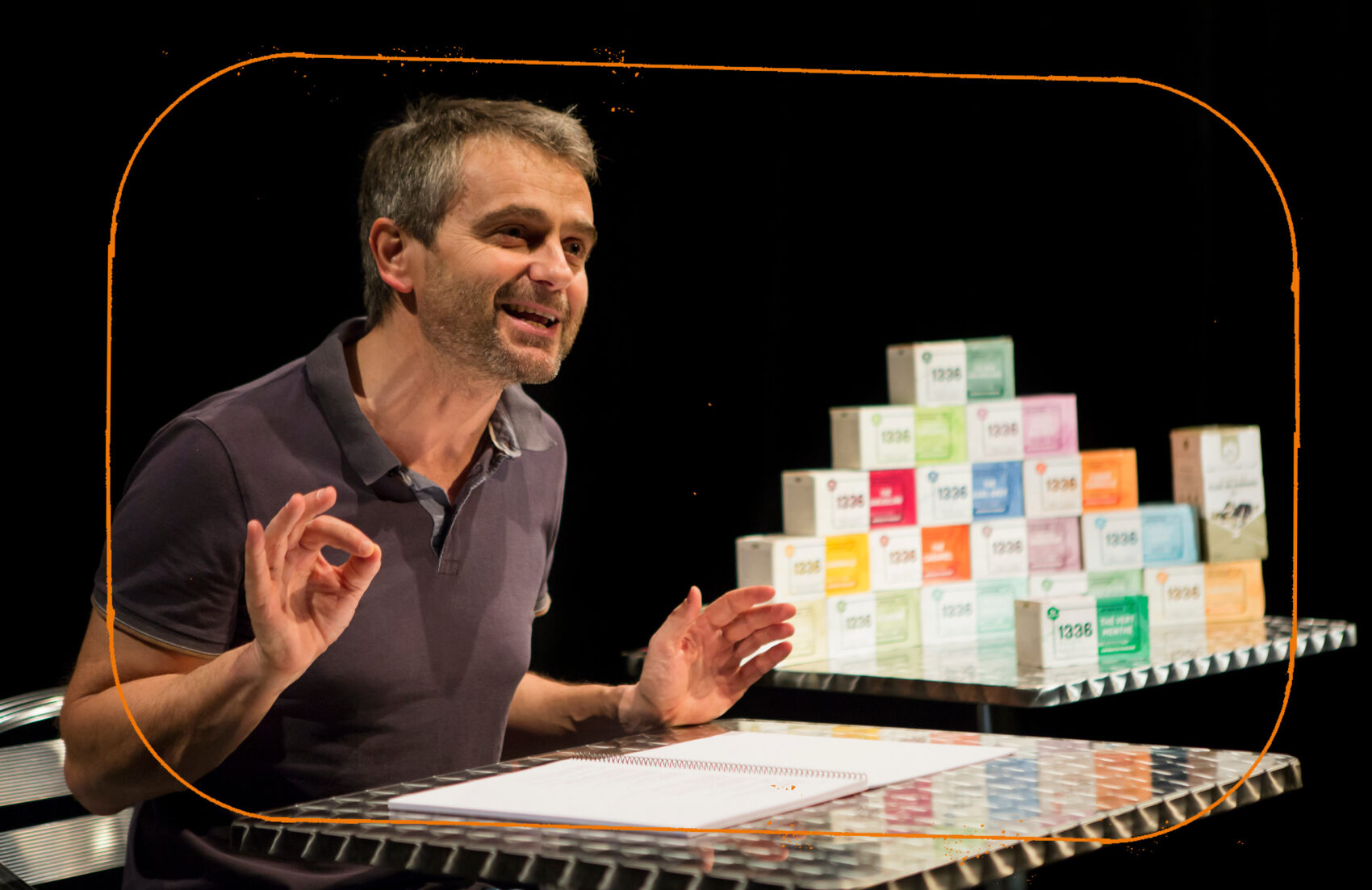 Photo de Philippe Durand sur scène assis devant une table avec un cahier. Avec derrière lui, une pyramide de boîtes de thé