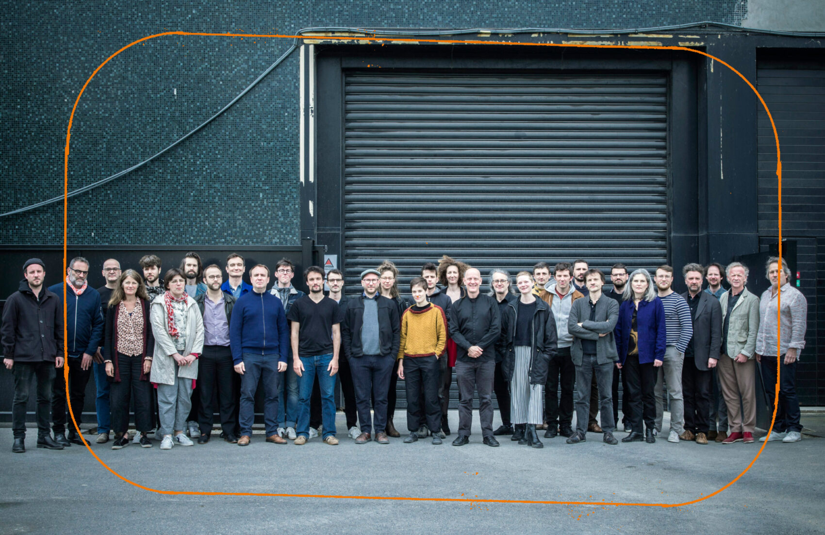 Tous les musiciens et musiciennes sont alignés pour faire une photo de classe, ils sont devant un grand hangar. 