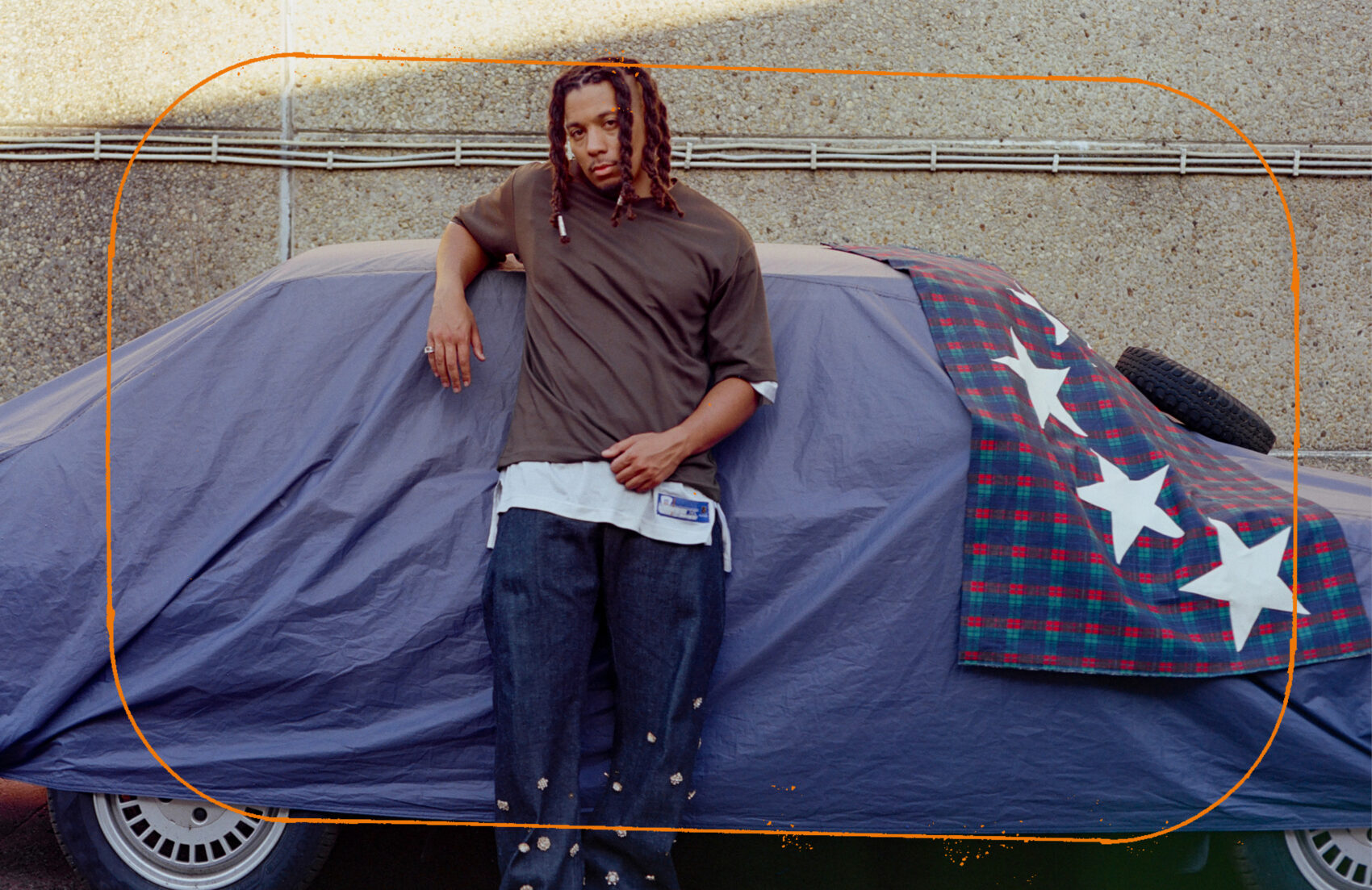 Photo du rappeur Swing accoudé à une voiture recouverte d'une bâche bleu