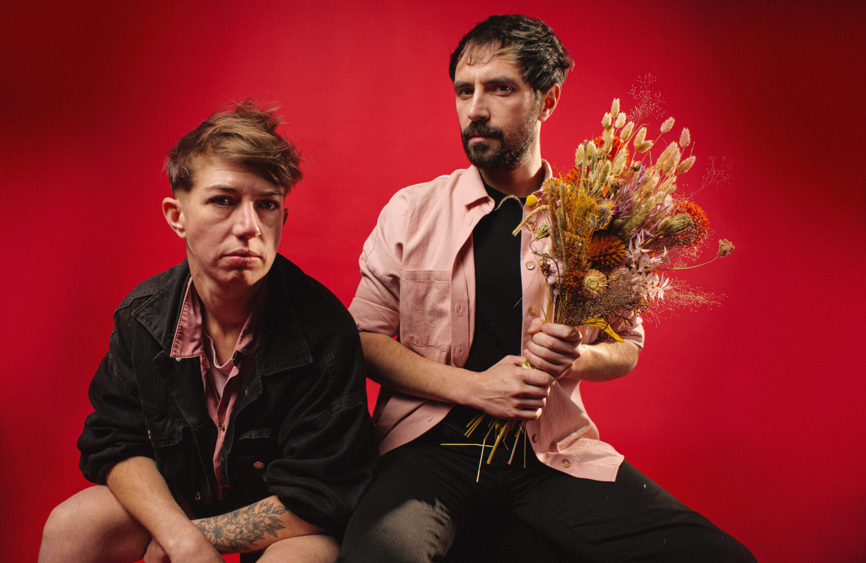 Photo du groupe La Ciguë avec un fond rouge, une femme gauche et un homme à droite avec un bouquet de fleurs séchées