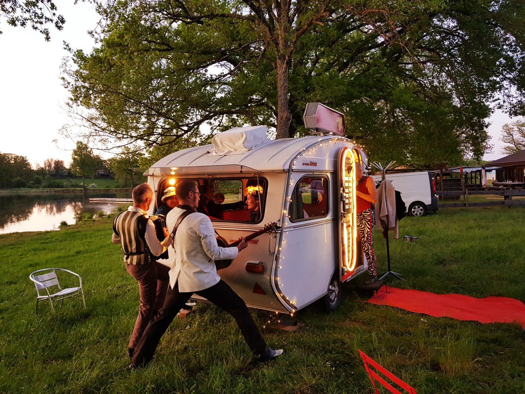 Une petite caravane avec des gens dedans et deux hommes à l'extérieur qui joue de la guitare face à la fenêtre
